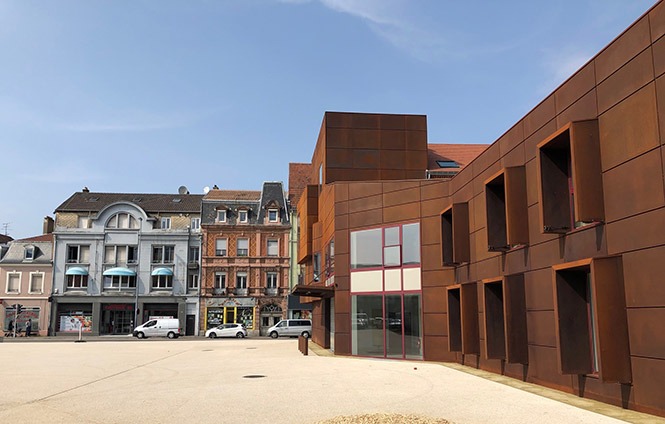 L'image montre une place publique entourée de bpatiment de maximum 2 étages, avec au premier plan un bâtiment en bois 'un étage