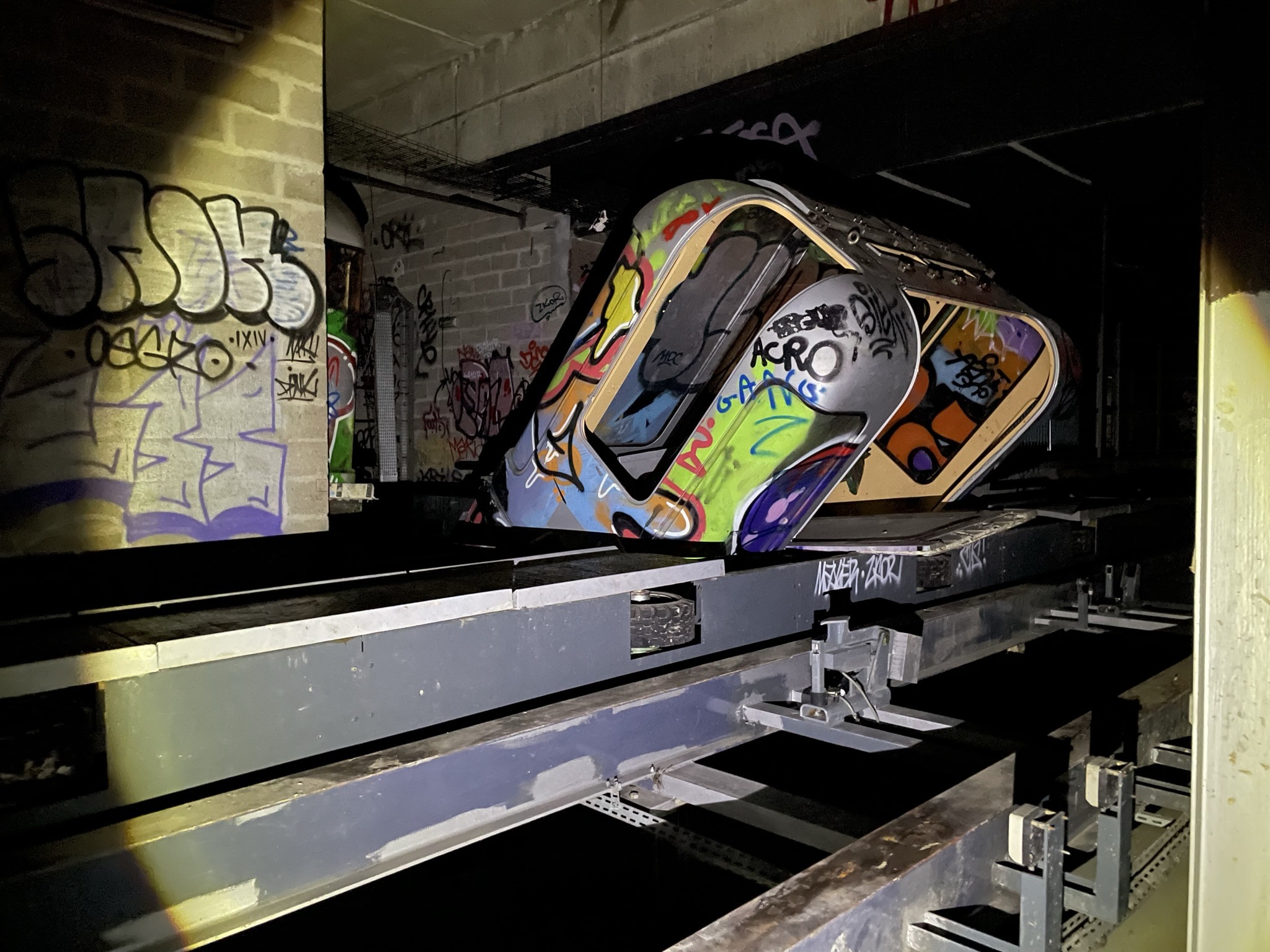 L'image montre un sous terrain tagué éclairé à la lampe, avec des rails et une rame de métro inclinée et taguée