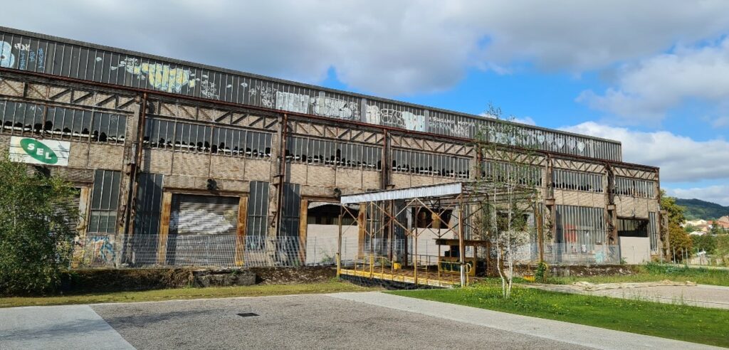 L'image montre une usine désaffectée en forme de hangar, avec des quais de déchargement pour camions recouverts de grafitis. Les ouvertures sont en mauvais état et les fenêtres cassées. Une place de parking au premier plan, du gazon, et une colline derrière
