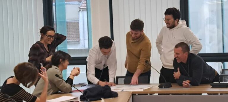 L'image montre 8 personnes assis et debout autour d'une table, ils sourient et montrent un plan