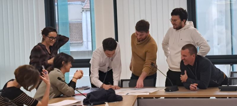 L'image montre 8 personnes assis et debout autour d'une table, ils sourient et montrent un plan