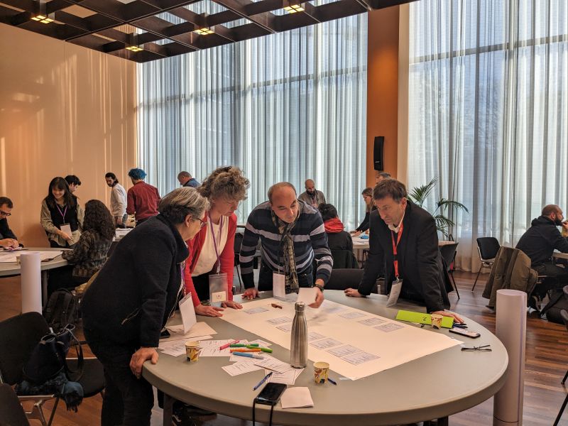 L'image montre 4 personnes debout autour d'une table sur laquelle est posée un plan