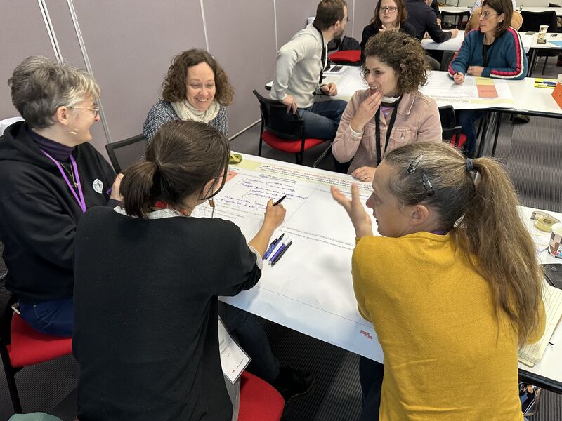 L'image montre 4 personnes assises autour d'une table, écrivant sur un grand tableau papier posé sur la table. elles sourient