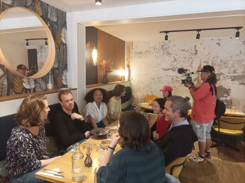 L'image montre 8 personnes attablées au restaurant, dont une se faisant interviewer par un cameraman et une speakeuse