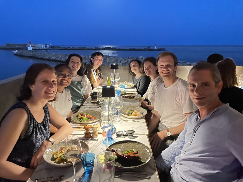 L'image montre 8 personnes souriantes assises autour d'une table dressée, devant la mer
