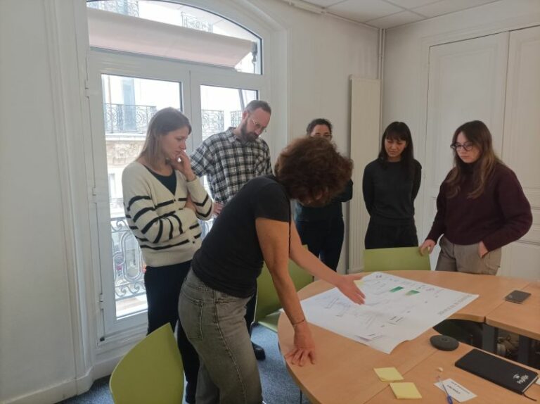 L'image montre 6 personnes dans un bureau autour d'une table, ils regardent un plan